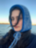 Woman wearing a blue knit bonnet by a calm seaside. Long dark hair flows in the breeze, creating a serene and reflective mood.