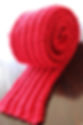 Bright red knitted scarf rolled up on a dark wooden surface, showcasing its textured pattern. The setting is simple, emphasizing the vibrant color.
