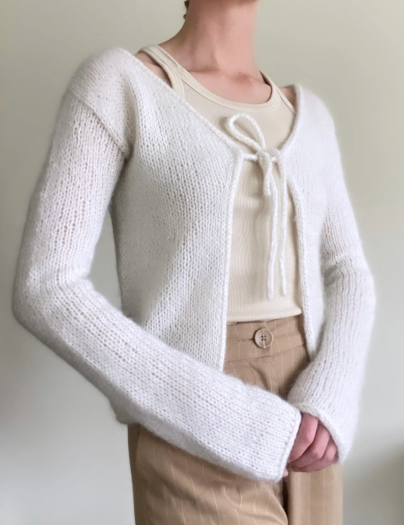 Person in a white knit cardigan over a beige top and tan pants. The cardigan is tied in front. Neutral background, soft lighting.