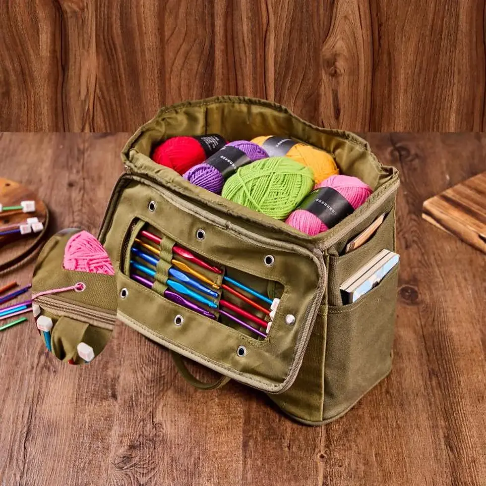 A khaki craft bag with colorful yarn and knitting needles on a wooden surface. A side pocket holds books, creating a cozy, organized setup.