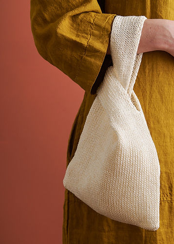 Person holding a textured white knit bag, wearing a mustard-yellow sleeve, set against a rust-colored background.