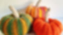 Three knitted pumpkins in green, orange, and multicolor sit on a white background. The setting is bright and cheerful with a cozy vibe.