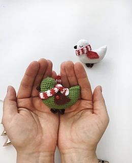 Hands holding a green crocheted bird wearing a red-striped scarf; another white bird with a similar scarf is in the background on a white surface.