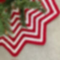 Red and white zigzag crochet tree skirt under a Christmas tree on a light carpeted floor. Green branches enhance the festive feel.