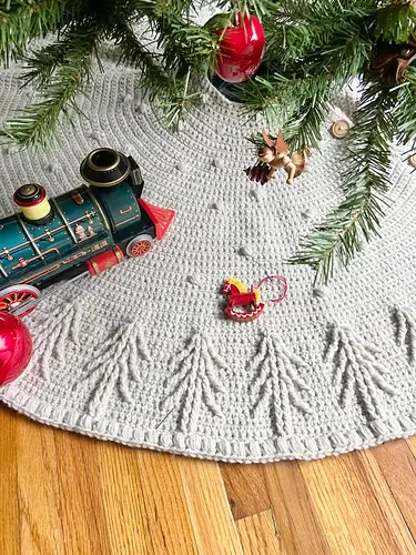 Toy train and reindeer on a crocheted tree skirt under a Christmas tree with red ornaments, set on a wooden floor, creating a festive scene.