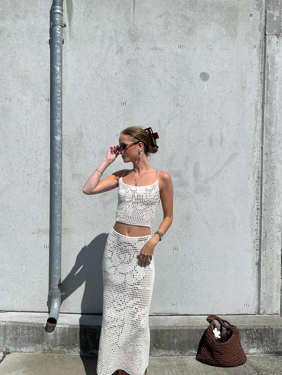 Woman in white crochet top and skirt adjusts sunglasses by a concrete wall. Brown bag on the ground; sunny day with calm mood.
