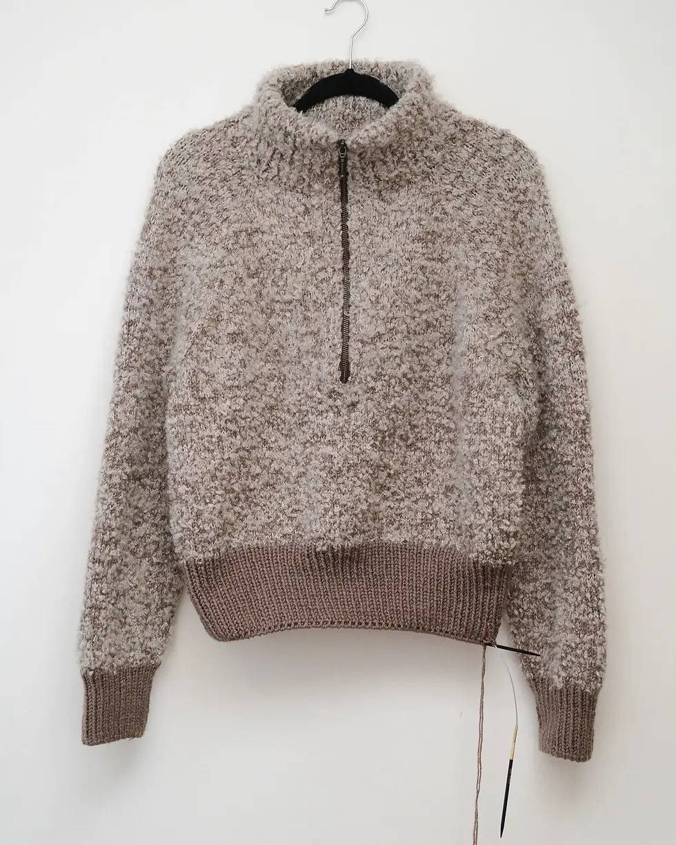 A fluffy beige sweater with a half-zip on a hanger against a white wall. Unfinished knitting work with needles dangles from the hem.