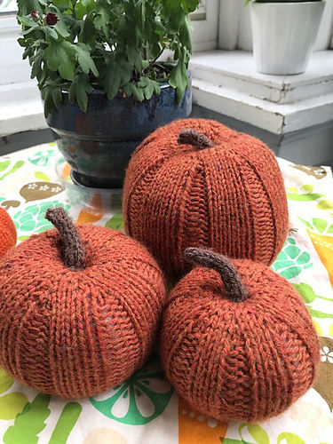 Knitted orange pumpkins on a patterned tablecloth, with a potted plant in the background. Cozy and autumnal setting.