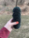 Hand holding a crocheted dark green plush with eyes, outdoors on grass. The hand has red nail polish, and a red sleeve is visible.