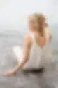 Woman in white knit top with back buttons sits on rock by water, looking relaxed. Light colors and serene mood.