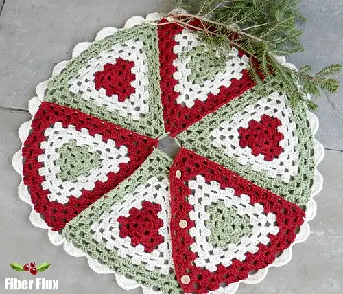Crocheted tree skirt with red, green, and white granny square pattern. Pine branches at the top; "Fiber Flux" logo at the bottom left.