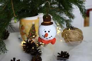 Crocheted snowman ornament hangs on pine branch, surrounded by pinecones. Background shows a festive mug and ball of twine. Cozy setting.