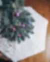 Christmas tree with purple and red ornaments stands on a white faux fur crochet tree skirt. The wooden floor background adds warmth to the festive scene.