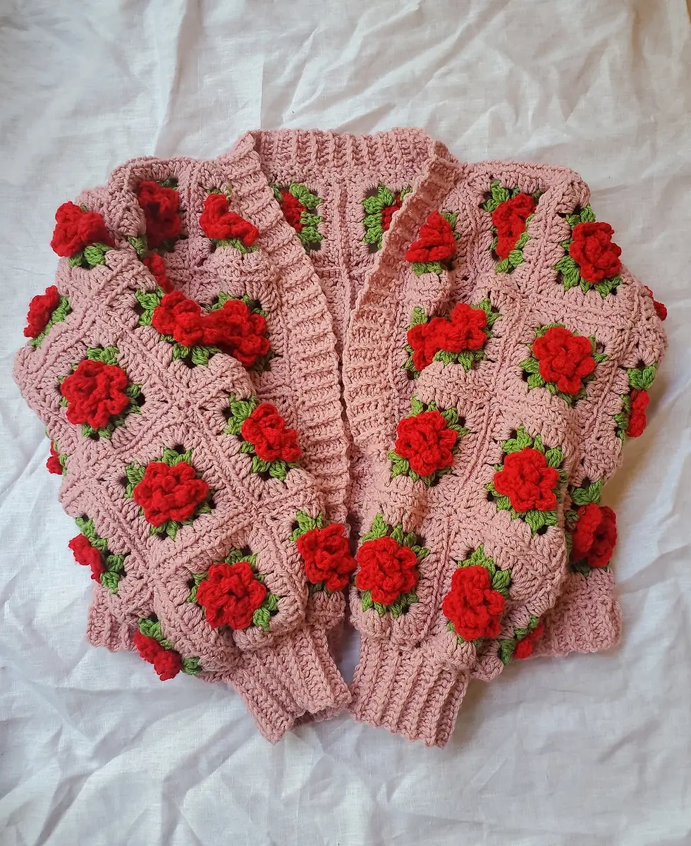 Pink crochet cardigan with red floral patterns and green leaves, laid flat on a crumpled white fabric background. Vibrant and cozy.