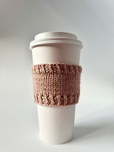 A white coffee cup with a pink knitted sleeve on a neutral background. The cup has a lid, and the setting is simple and minimalist.