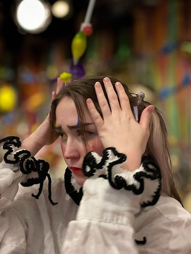 Person with hands on head in a colorful, whimsical setting. Wearing a white top with black patterns, expression appears thoughtful.