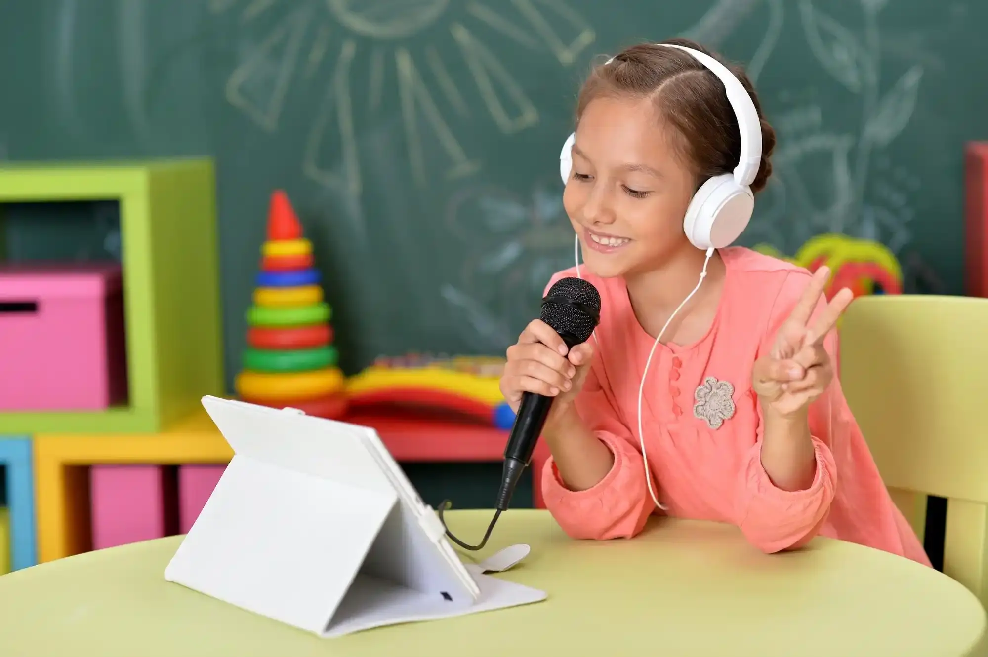 Smiling girl in headphones sings into microphone, engaging with tablet.