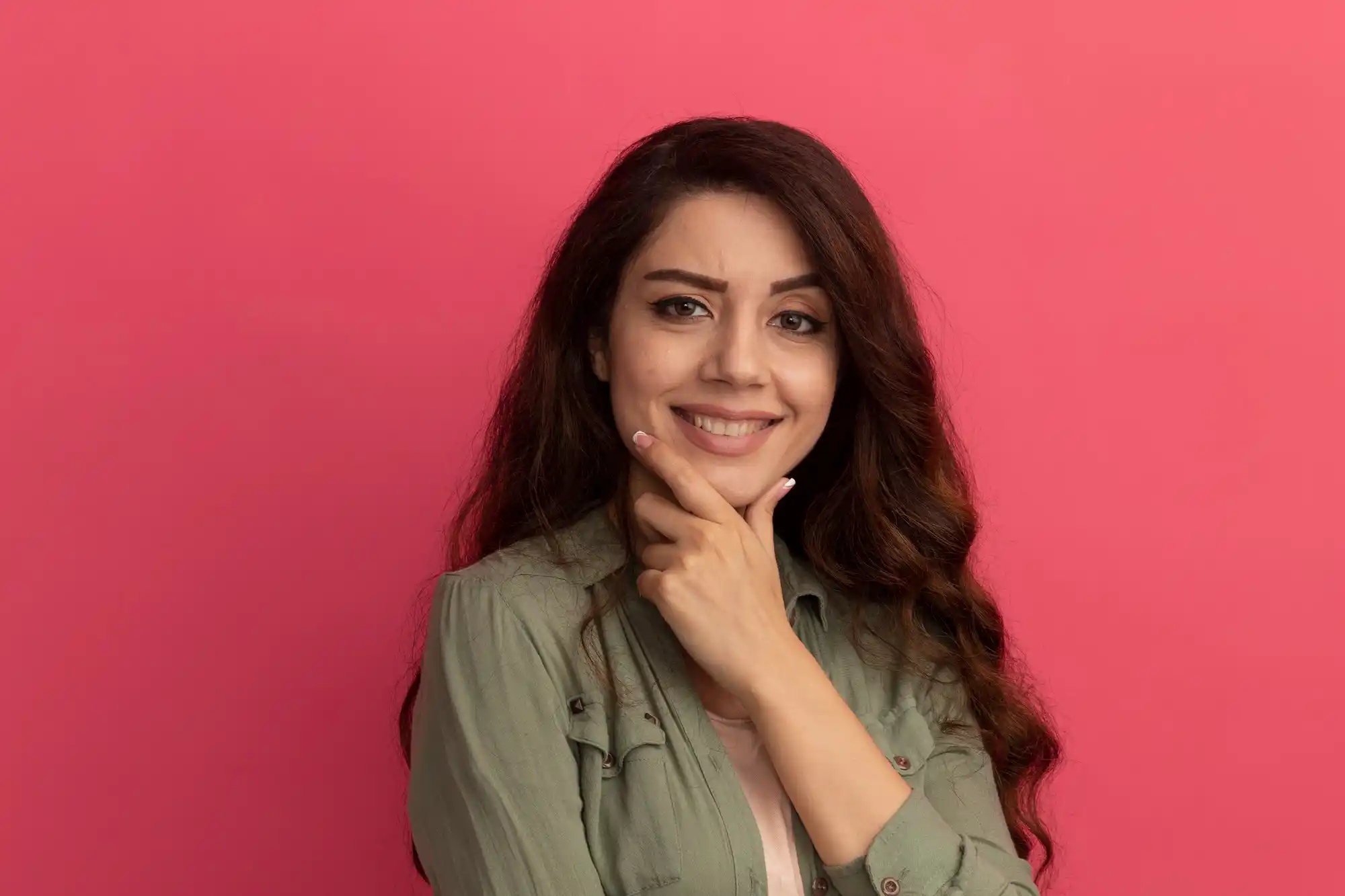 Smiling young woman, long brown hair, hand on chin, pink background.