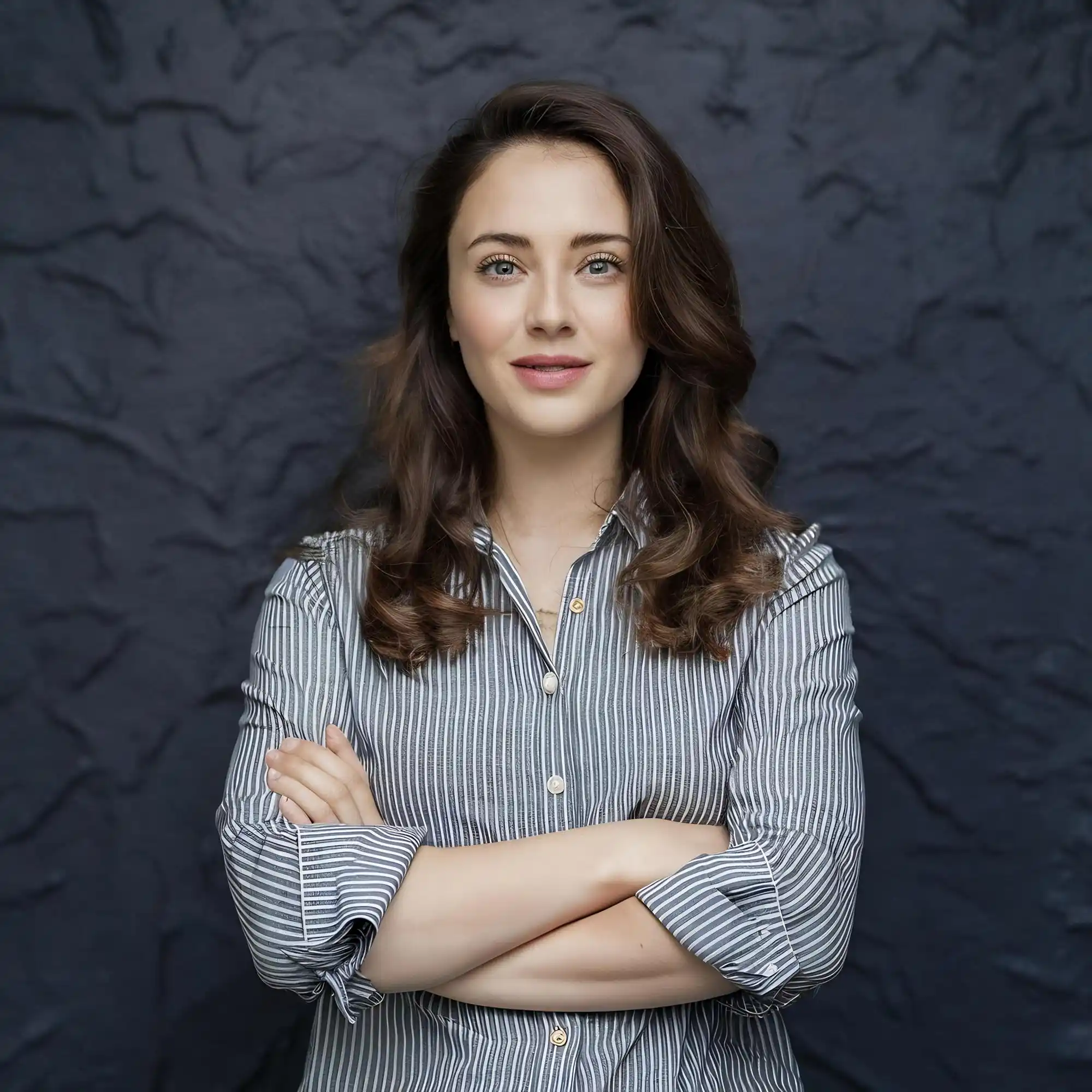Smiling woman with wavy brown hair in grey striped shirt, arms crossed.