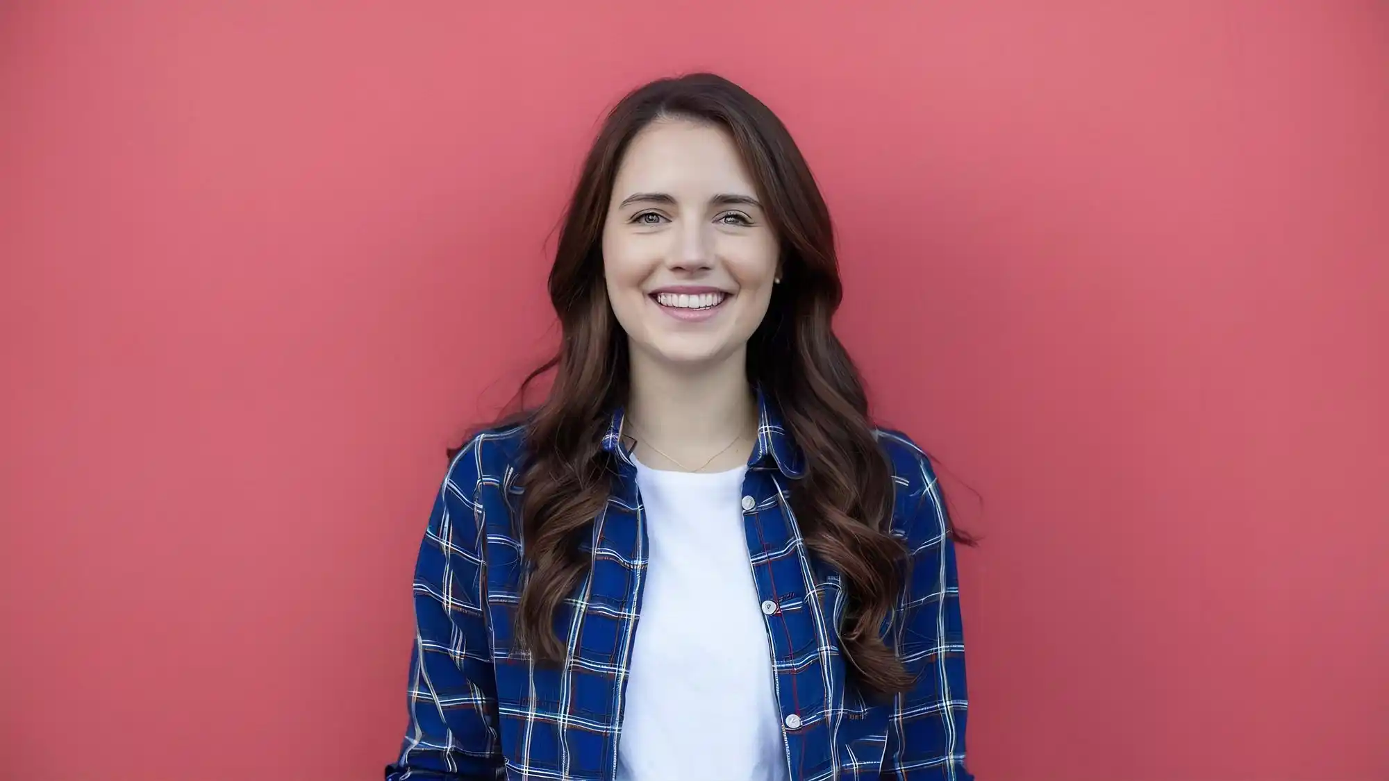 Young woman smiling, wearing a blue plaid shirt against a red wall.