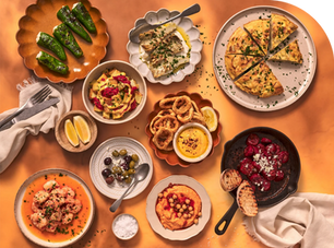 Top-down spread of colourful tapas dishes including: padron peppers, dips, olives, garlic prawns, tortilla and chorizo on an orange table.