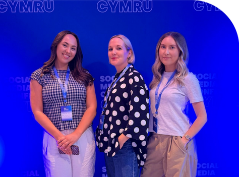 Three women wearing lanyards pose smiling at a Social Media Conference Cymru backdrop with blue lighting.