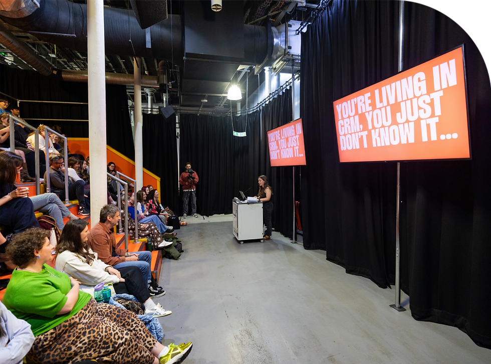 Audience seated in a seminar room watching a speaker. Screen displays: "YOU'RE LIVING IN CRM, YOU JUST DON'T KNOW IT..." Black curtains backdrop.