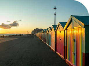 Colourful beach huts line Brighton promenade at sunset. People walk in the distance, with a calm sea to the left and a clear sky above.