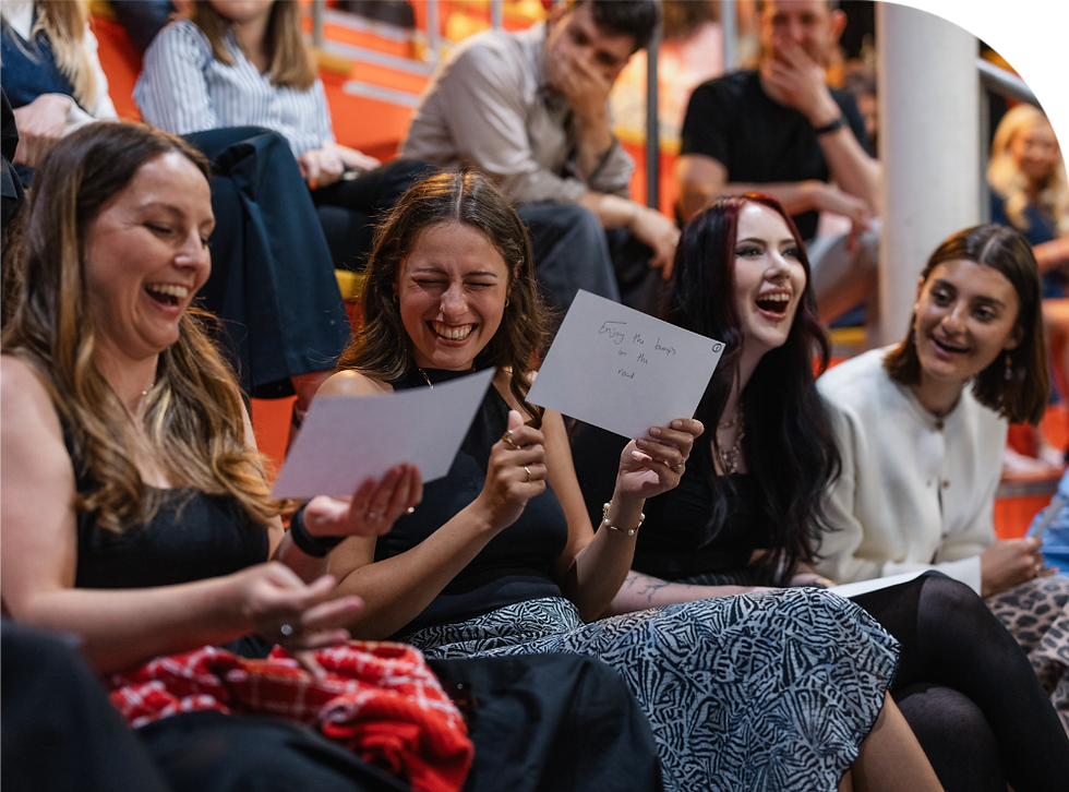 Four women sit and laugh, holding papers with text. They're surrounded by a crowd in an auditorium setting. The mood is joyful and vibrant.