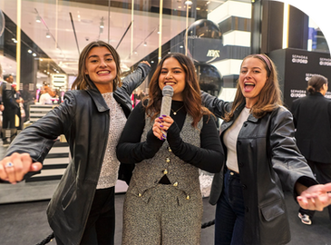 Three women smiling and posing with a microphone in a lively indoor setting. There are Sephora signs in the background.