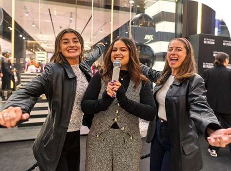 Three women smiling and posing with a microphone in a lively indoor setting. There are Sephora signs in the background.