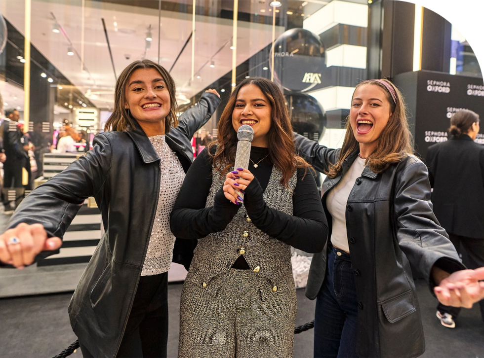 Three women smiling and posing with a microphone in a lively indoor setting. There are Sephora signs in the background.