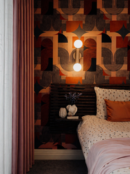 A luxurious contemporary bedroom featuring a striking geometric feature wall in rich terracotta, black, and bronze tones, creating a dramatic focal point behind the bed. A sculptural sand wall light with globe bulbs provides warm ambient lighting, while blush pink curtains, layered bedding, and textured cushions soften the bold design. A floating bedside shelf and carefully curated accessories complete the elegant, boutique hotel-inspired interior.