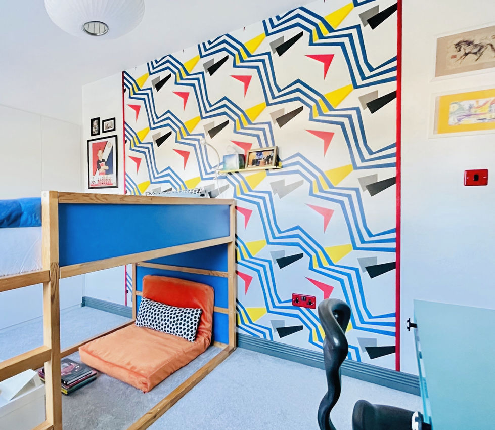 A contemporary children's bedroom featuring a bespoke wooden cabin-style loft bed with bold blue panels, set against a vibrant geometric feature wall in shades of blue, mustard yellow, coral red, black, and white. A cosy floor cushion beneath the bed creates a relaxing reading nook, while floating shelves, framed artwork, and playful accessories add personality. The bright colours, graphic patterns, and space-saving design combine to create a fun, modern bedroom for a growing child.