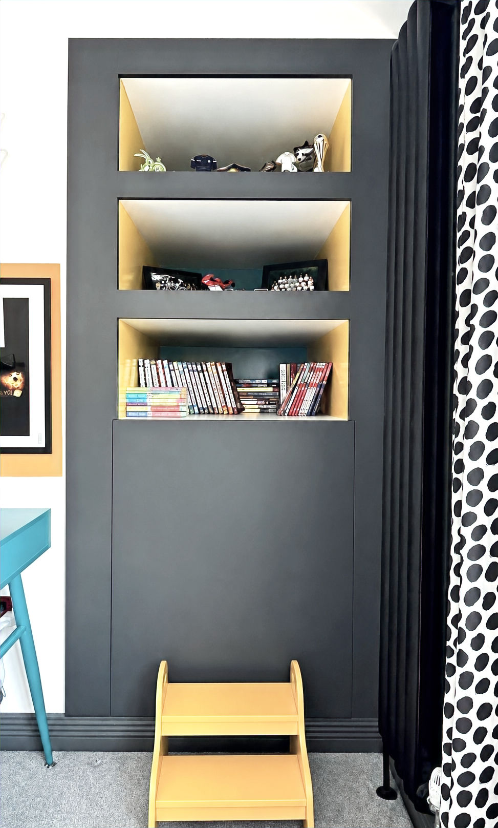 A bespoke charcoal-grey display cabinet with integrated warm LED lighting provides stylish storage for books, model cars, and collectables in a contemporary children's bedroom. A light wood step stool offers easy access to the lower shelves, while black-and-white spotted curtains and a bright blue desk add playful contrast. The built-in joinery combines practical storage with a clean, modern aesthetic.