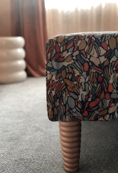 A close-up of an upholstered bedroom bench featuring a richly patterned fabric in terracotta, blush, charcoal, and muted blue tones, paired with turned timber legs. Soft natural light, plush carpeting, and blurred blush curtains in the background create a warm, inviting atmosphere, while the textured fabric adds colour and visual interest to the contemporary bedroom design.