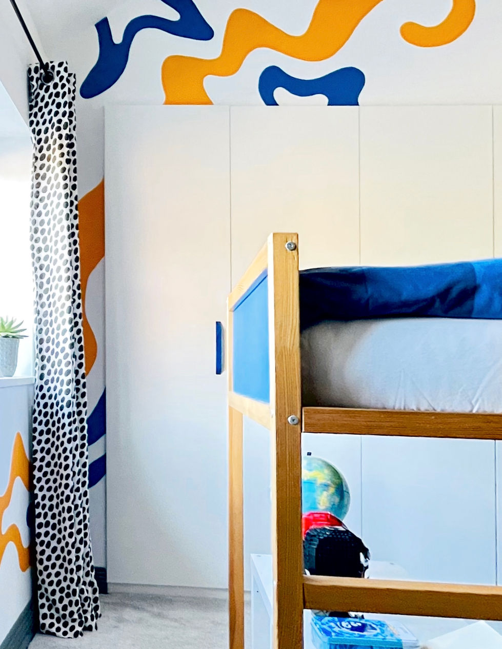 A contemporary children's bedroom featuring a wooden cabin-style loft bed alongside bespoke white fitted wardrobes. A bold abstract mural in vibrant blue and mustard yellow flows across the walls and ceiling, creating a playful statement. Black-and-white spotted curtains add graphic contrast, while the bright colour palette and clever space planning combine to create a fun, modern room designed for both sleep and play.