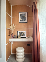 A contemporary dressing area featuring warm natural wood cladded wall panels with crisp white trim, creating a refined geometric backdrop. A floating white dressing table is styled with decorative accessories, framed abstract artwork, and a sculptural cream stool, while blush pink curtains soften the space with warmth and texture. The thoughtfully layered finishes and calming colour palette create an elegant, boutique-inspired dressing nook.