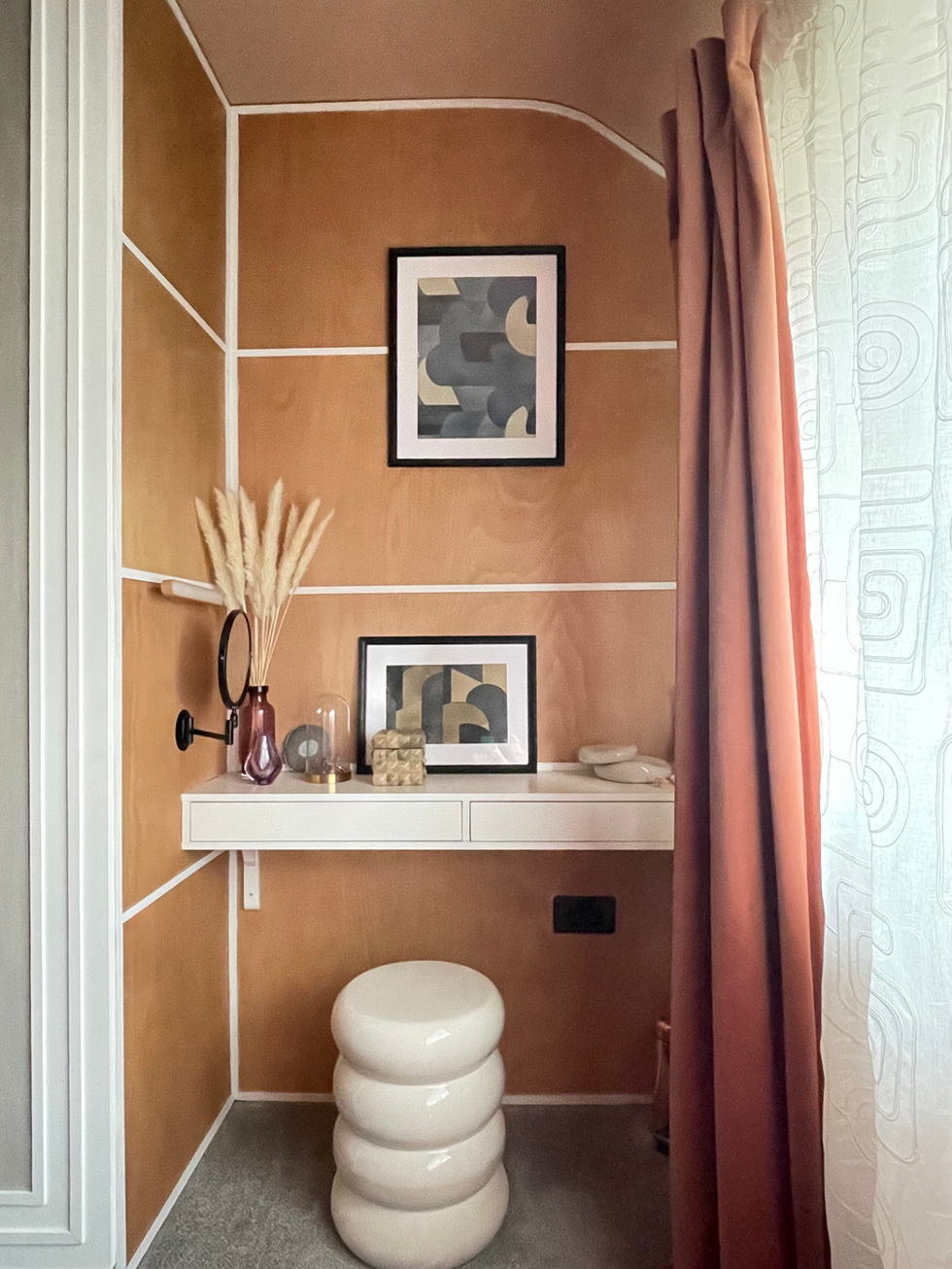 A contemporary dressing area featuring warm natural wood cladded wall panels with crisp white trim, creating a refined geometric backdrop. A floating white dressing table is styled with decorative accessories, framed abstract artwork, and a sculptural cream stool, while blush pink curtains soften the space with warmth and texture. The thoughtfully layered finishes and calming colour palette create an elegant, boutique-inspired dressing nook.