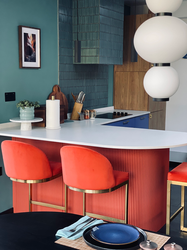 A contemporary kitchen island featuring a crisp white worktop and a fluted coral-red breakfast bar, paired with upholstered coral bar stools with brushed brass bases. Rich teal wall tiles, warm wood cabinetry, and vibrant cobalt blue base units create a bold, layered colour palette, while sculptural globe pendant lights add a striking focal point. The bespoke design combines colour, texture, and functionality to create an inviting space for cooking, dining, and entertaining.