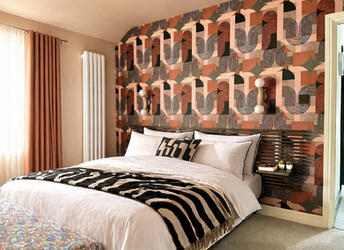 A luxurious contemporary bedroom featuring a dramatic geometric feature wall in terracotta, blush pink, charcoal, and black tones behind the bed. Crisp white bedding is layered with bold monochrome patterned cushions and a zebra-inspired throw, while sculptural wall lights and floating slatted bedside shelves add warmth and sophistication. Soft blush curtains, a vertical designer radiator, and carefully curated accessories complete this elegant, boutique hotel-inspired interior.