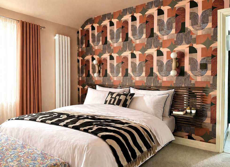 A luxurious contemporary bedroom featuring a dramatic geometric feature wall in terracotta, blush pink, charcoal, and black tones behind the bed. Crisp white bedding is layered with bold monochrome patterned cushions and a zebra-inspired throw, while sculptural wall lights and floating slatted bedside shelves add warmth and sophistication. Soft blush curtains, a vertical designer radiator, and carefully curated accessories complete this elegant, boutique hotel-inspired interior.