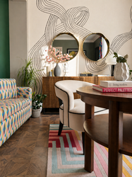 A contemporary reading area featuring a striking black-and-white mural with flowing abstract linework, complemented by two round brass-framed mirrors above a cane-front sideboard. A round mid-century coffee table is paired with elegant upholstered chairs and a colourful geometric rug, while a patterned accent chair, indoor plants, and carefully styled accessories add warmth and personality. Parquet wood flooring and layered textures create a sophisticated, design-led space for cap of tea and a good book.