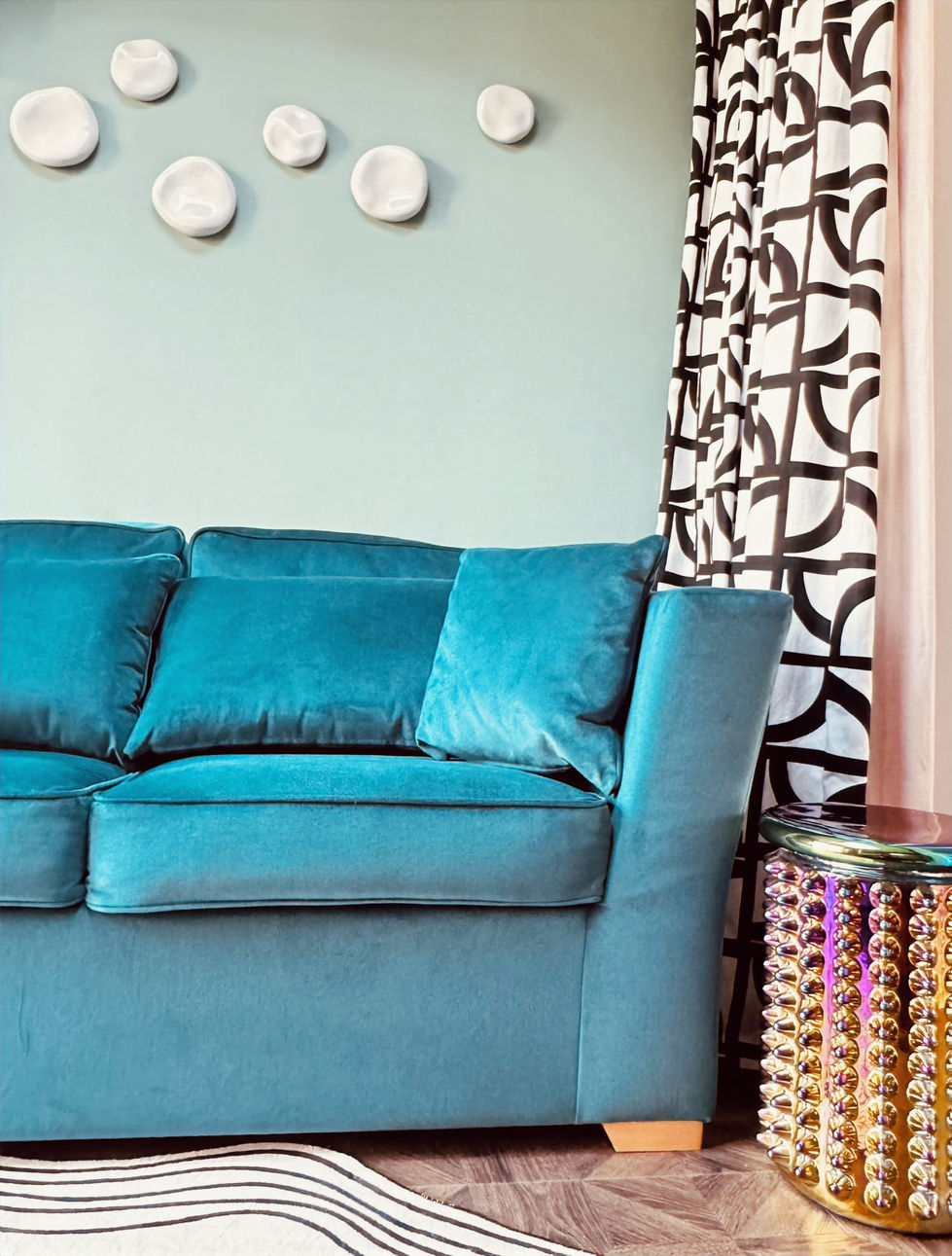 A contemporary guest room featuring a luxurious teal velvet sofa set against a soft sage green wall, accented with sculptural white ceramic wall art. Bold black-and-white patterned curtains frame the space, while a metallic side table with an iridescent beaded finish adds texture and glamour. Herringbone wood flooring and a monochrome abstract rug complete the sophisticated, modern interior.