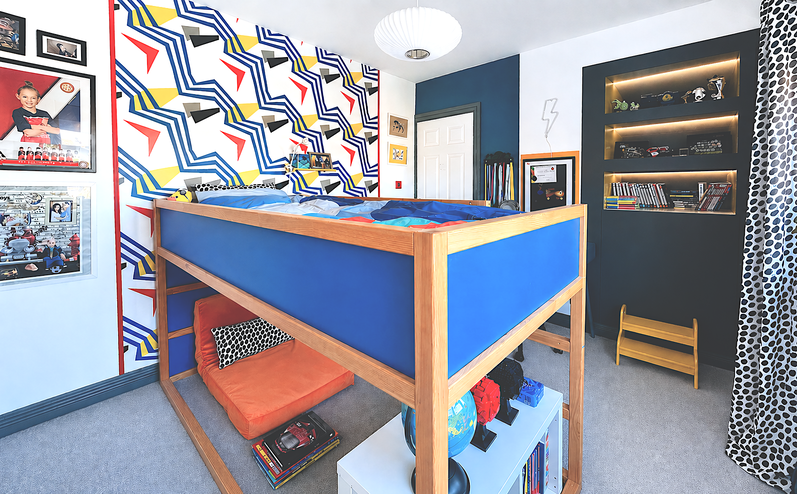 A contemporary children's bedroom featuring a bespoke wooden cabin-style loft bed with bold blue panels, creating a multifunctional space for sleep, play, and study. A striking geometric feature wall in blue, mustard yellow, black, red, and white adds energy and personality, while illuminated charcoal-grey display shelving showcases books and collectables. Coordinated blue walls, playful accessories, and clever built-in storage complete this vibrant, modern family interior.