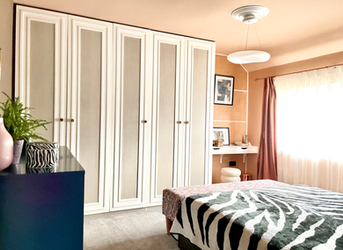 A luxurious contemporary bedroom featuring bespoke floor-to-ceiling wardrobes with elegant panelled doors, complemented by warm terracotta walls with crisp white detailing. Soft blush curtains frame a large window, filling the room with natural light, while a cosy bed dressed in crisp white bedding and a bold zebra-inspired throw adds contrast and texture. In the corner, a stylish dressing nook with a floating vanity and sculptural stool enhances the boutique hotel-inspired feel of the space.
