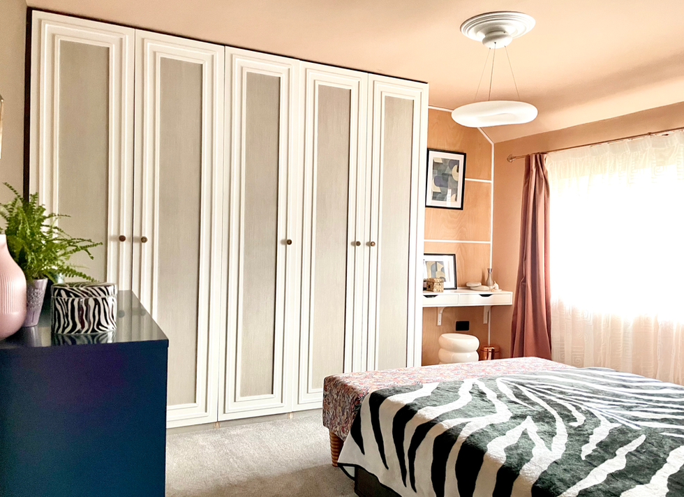 A luxurious contemporary bedroom featuring bespoke floor-to-ceiling wardrobes with elegant panelled doors, complemented by warm terracotta walls with crisp white detailing. Soft blush curtains frame a large window, filling the room with natural light, while a cosy bed dressed in crisp white bedding and a bold zebra-inspired throw adds contrast and texture. In the corner, a stylish dressing nook with a floating vanity and sculptural stool enhances the boutique hotel-inspired feel of the space.