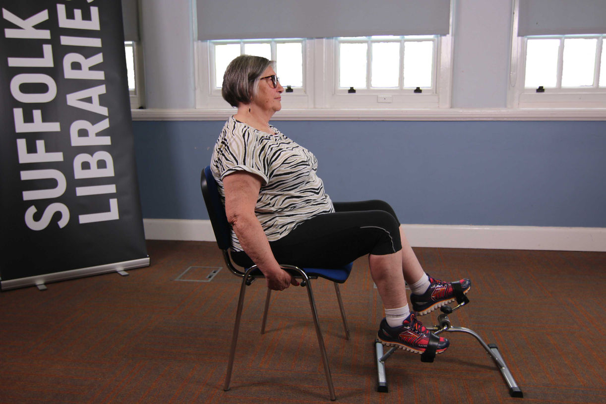 Wider shot of elderly lady demonstrating a seated excercise with portable pedalling exercise equipment.