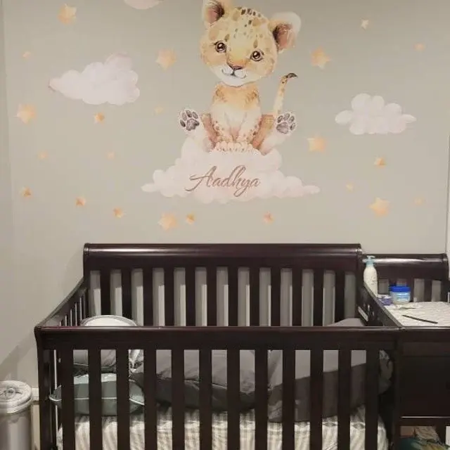 Ein personalisierter Kinderzimmer-Wandaufkleber mit einem Aquarell-Baby-Löwenjungen auf einer Wolke mit dem Wunschnamen 'Aadhya', umgeben von Sternen über einem Babybett.