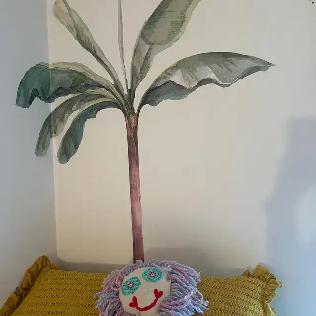 Ein großer Aquarell-Palmen-Wandaufkleber, der einem Kinderzimmer oder einem Boho-Kinderzimmer ein tropisches Dschungelthema verleiht, positioniert an einer Wand hinter einem gelben Kissen.